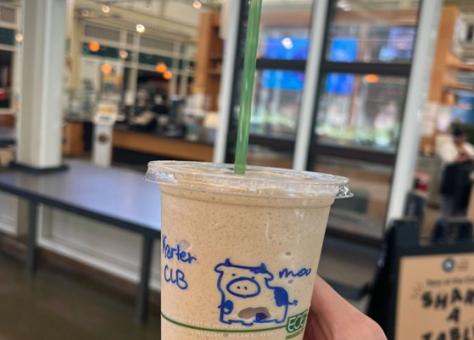 A hand holds a plastic cup filled with a blended iced drink, featuring a green straw and a drawing of a cow on the cup, inside a café with large windows.
