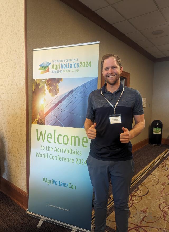 Jack Schaufler holding up two thumbs up, standing next to a welcome sign at a conference