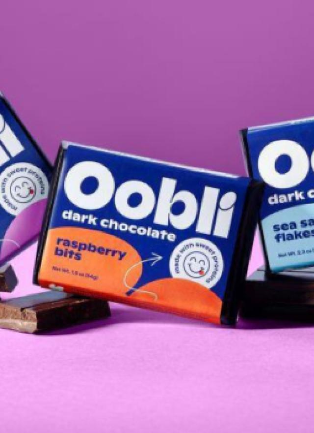 Oobli dark chocolate bars in Silky 70% Cacao, Raspberry Bits, and Sea Salt Flakes flavors displayed on chocolate squares against a purple background.
