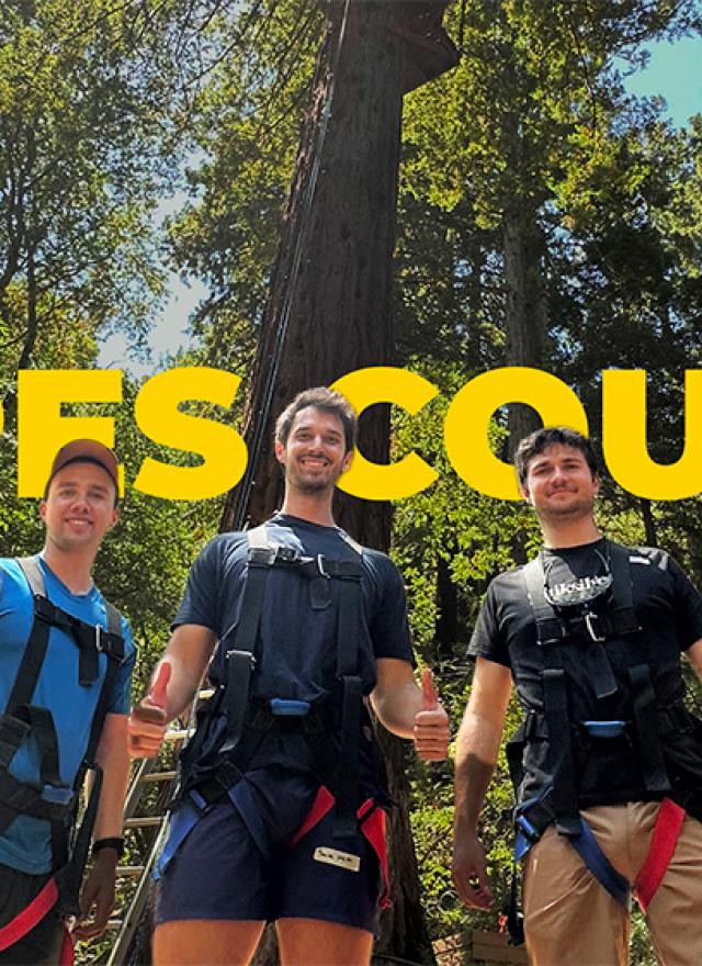 Three UC Davis MBA students wearing harnesses pose together at the Challenge Sonoma Ropes Course in Glen Ellen, California, standing in front of tall redwood trees with the words “ROPES COURSE” in bold yellow behind them.