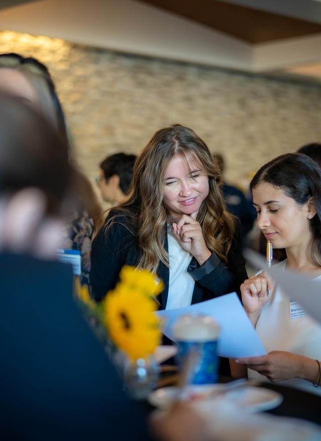 Two MBA students in professional attire review a document together at a networking event, surrounded by other attendees. A sunflower centerpiece and notepads are visible on the table.