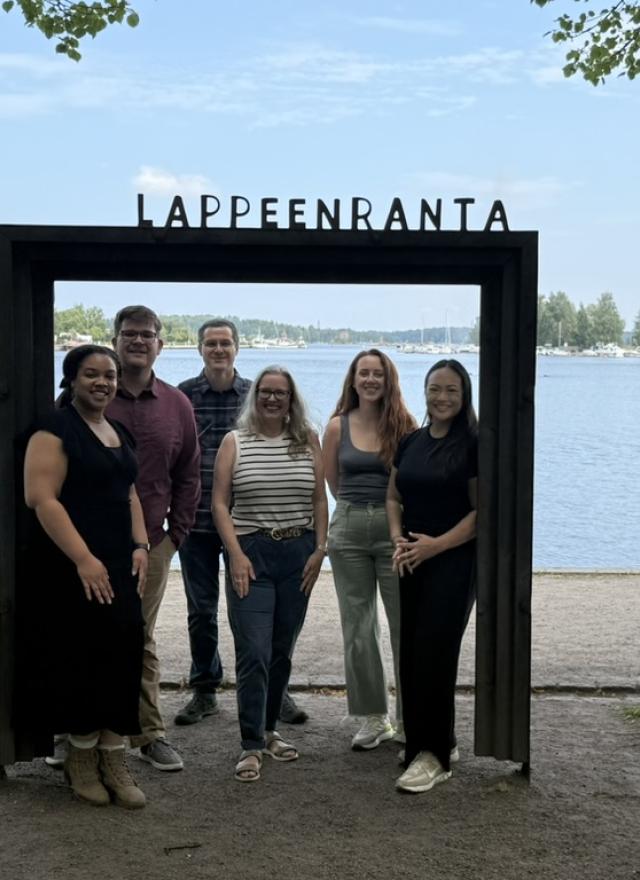 "Six people stand smiling inside a large black rectangular frame with the word 'Lappeenranta' on top. Behind them is a scenic lakeside view with boats, trees, and a partly cloudy sky