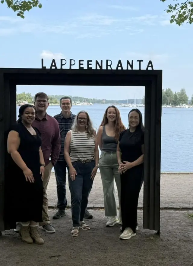 "Six people stand smiling inside a large black rectangular frame with the word 'Lappeenranta' on top. Behind them is a scenic lakeside view with boats, trees, and a partly cloudy sky