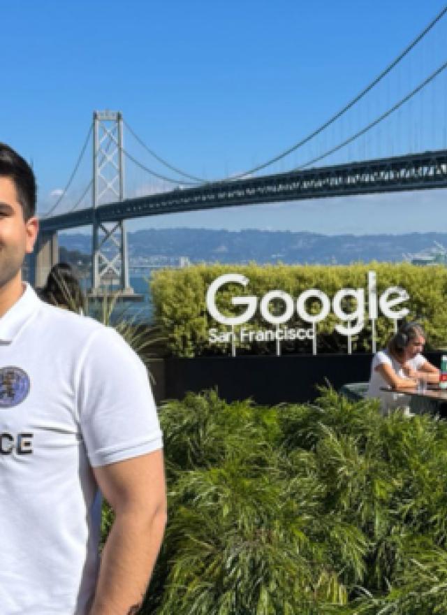 Talha Amin stands on an outdoor terrace at Google’s San Francisco office, with the Bay Bridge and blue sky in the background. He’s wearing a white polo shirt, light pants and sunglasses, surrounded by lush green plants.