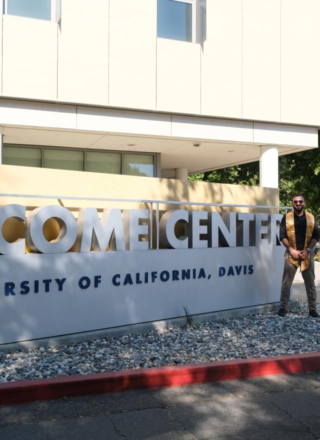 Sanjay Puri standing in front of UC Davis Welcome Center, wearing a graduation stole