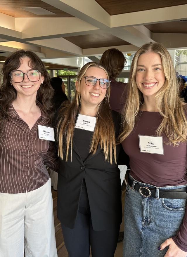 Three UC Davis MPAc students, including Mia Hildebrand on the right, stand together and smile at a networking event indoors.