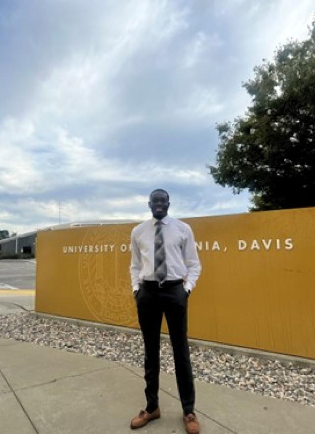 A man in formal attire stands in front of a yellow University of California, Davis sign under a partly cloudy sky.