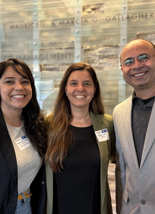 Three people stand indoors, smiling at the camera. They wear business attire and conference name tags. A wall sign behind them reads "Graduate School of Management.