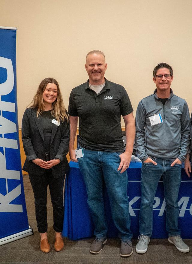 Four people stand and smile beside a KPMG banner and table in a conference room, wearing business casual attire and name tags.
