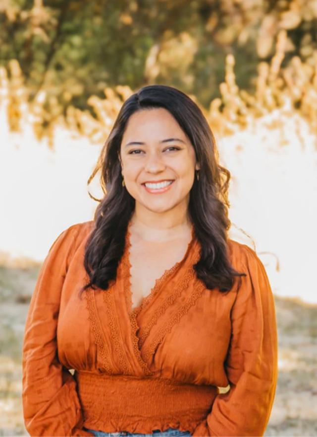 A woman with long dark hair, wearing an orange blouse, stands outdoors smiling, with blurred greenery and sunlight in the background.