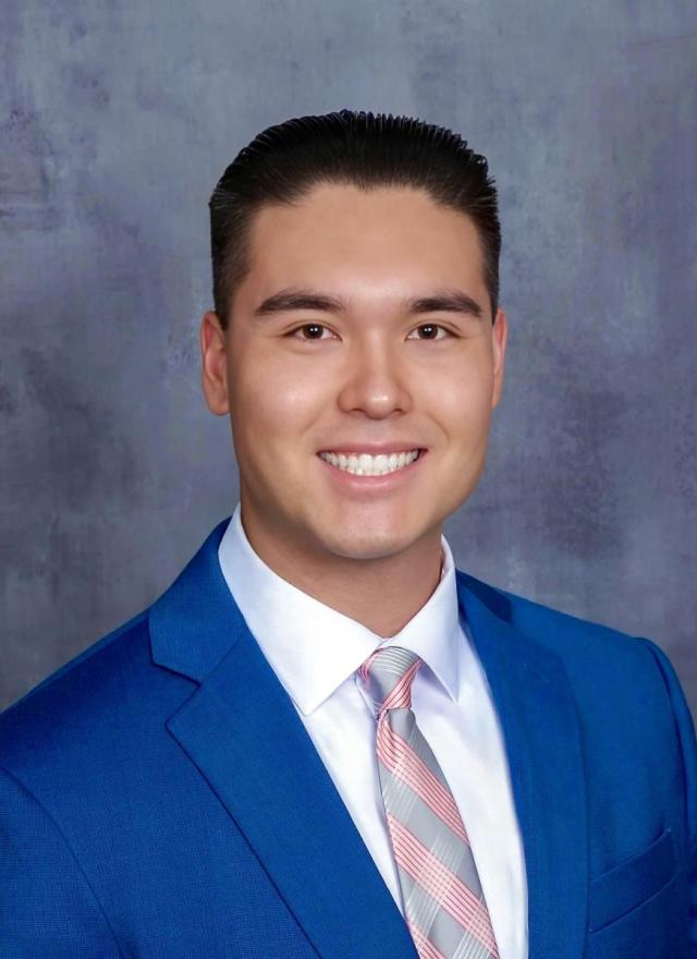 A young man wearing a blue suit, white shirt, and striped tie smiles in front of a gray mottled background.