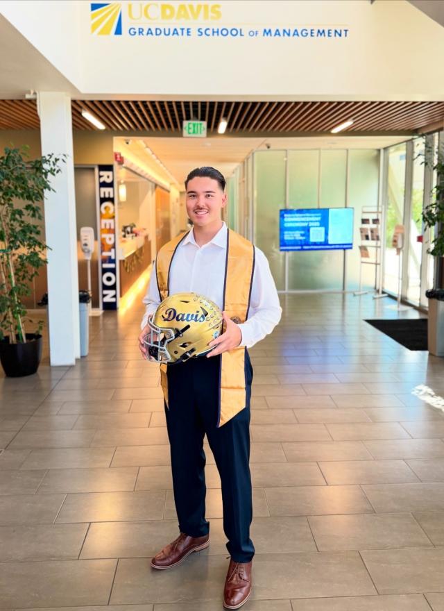 A man in business attire and a graduation stole stands holding a UC Davis football helmet in the lobby of the UC Davis Graduate School of Management.