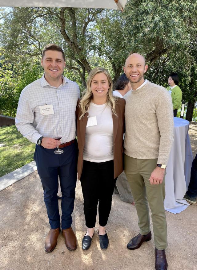 Three people stand outdoors, smiling at the camera. The two men wear business casual attire; the woman wears a white top and brown blazer. Trees and other event attendees are in the background.
