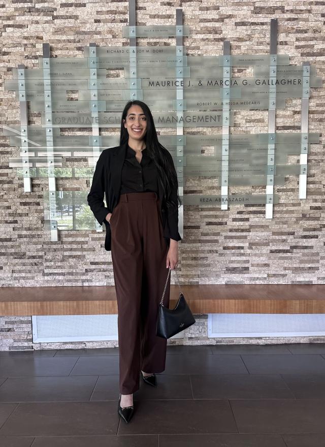 A woman stands indoors in business attire, holding a handbag and smiling, in front of a wall displaying names and a sign reading “Graduate School of Management.”