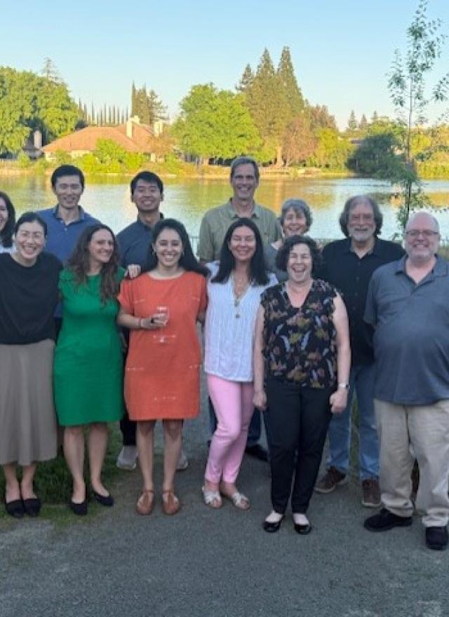 13 professors at the conference standing outdoors in front of a lake, smiling at the camera on a sunny day.