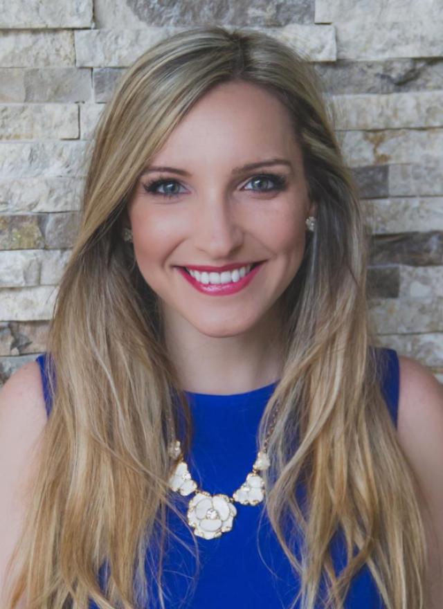 A woman with long blonde hair, wearing a blue sleeveless top and a floral necklace, smiles in front of a textured stone wall background.