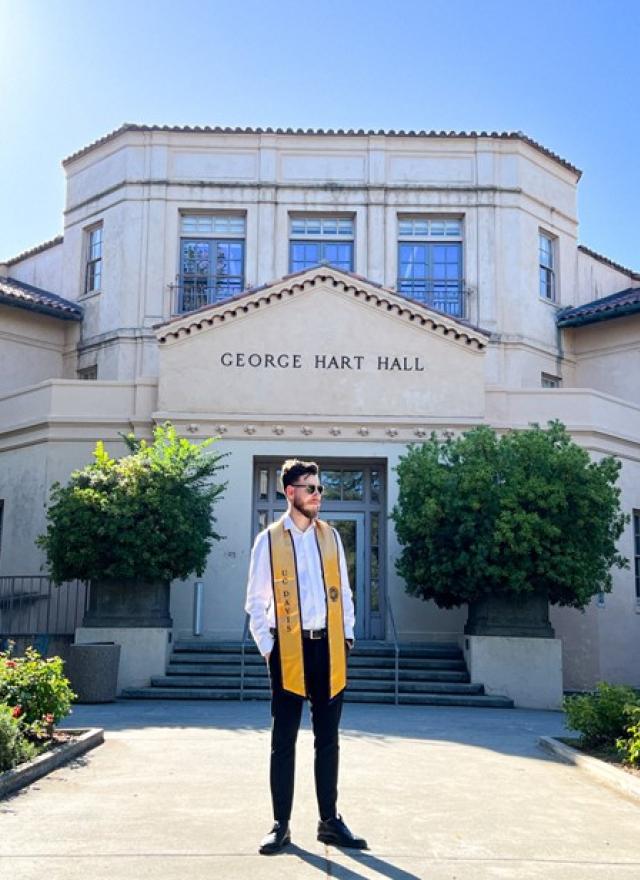 A man wearing a graduation stole stands in front of George Hart Hall on a sunny day.