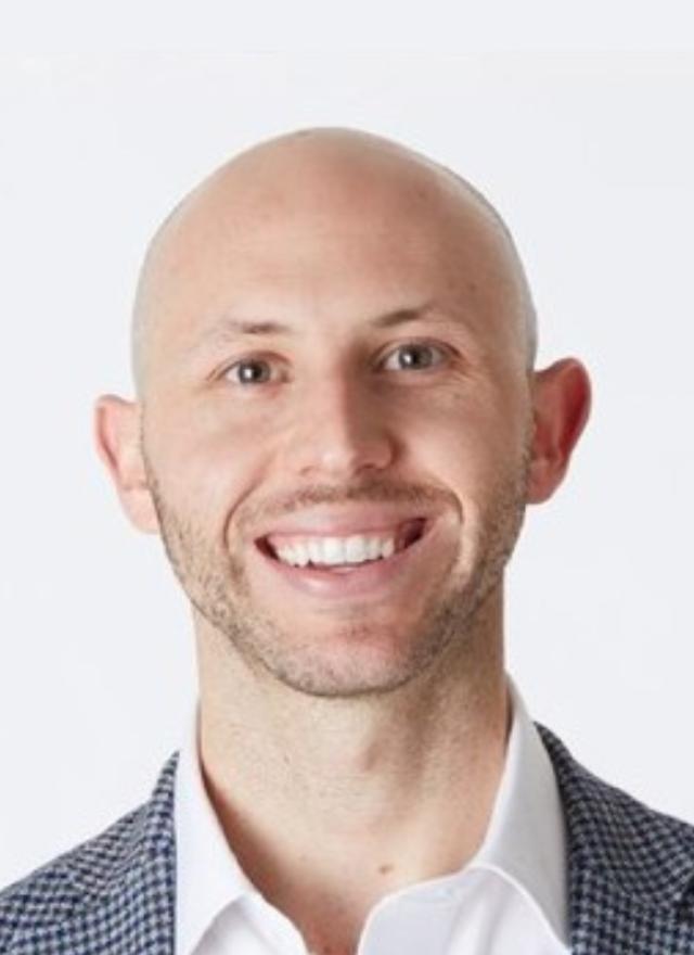 A smiling bald man wearing a white shirt and a checkered blazer, posed against a plain white background.