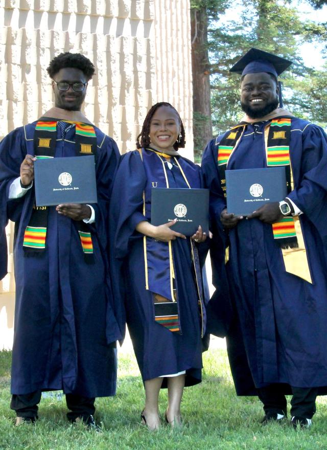 Three graduates in navy blue caps and gowns hold diplomas and smile outdoors; another person walks in the background.