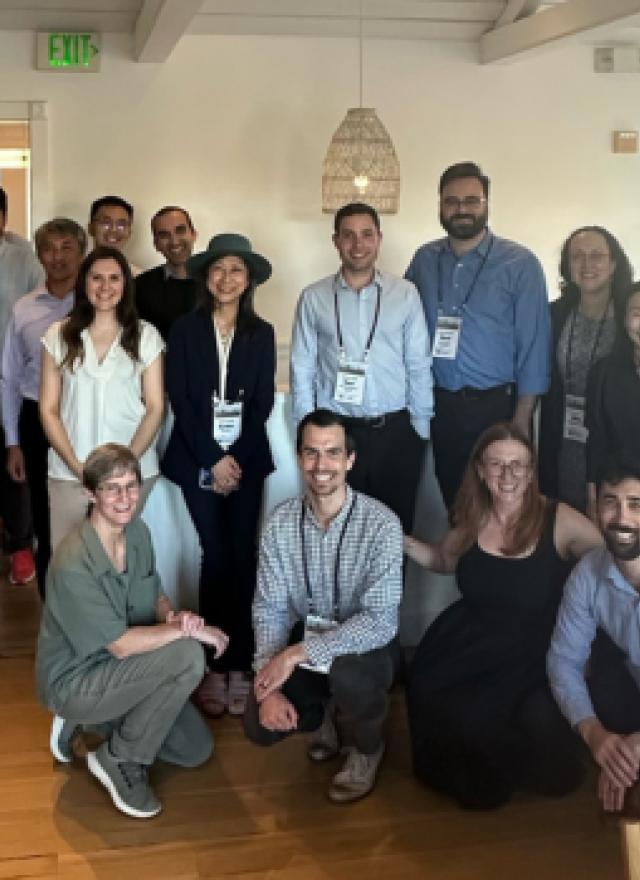 A group of about 20 people pose together indoors, some standing and some kneeling, in a well-lit room with wooden floors and white walls.