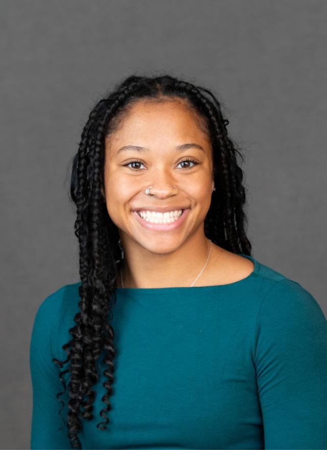 A woman with long braided hair, wearing a teal top, smiles at the camera against a plain gray background.
