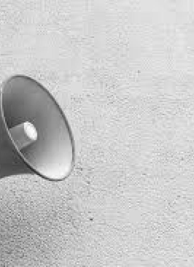 A wall-mounted megaphone or loudspeaker is shown against a plain, textured wall in black and white.