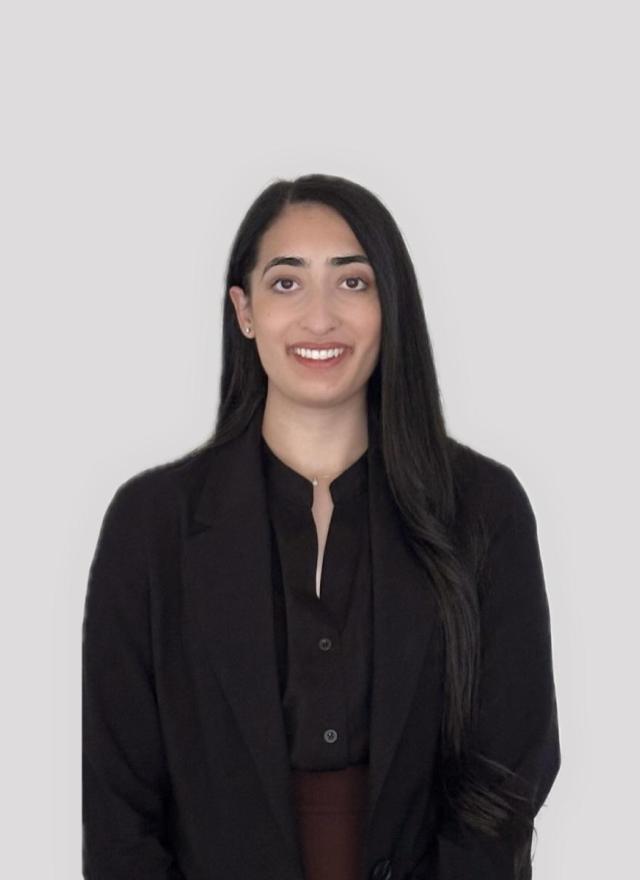 A woman with long dark hair, wearing a black blazer and dark shirt, stands in front of a plain light gray background, facing the camera and smiling.