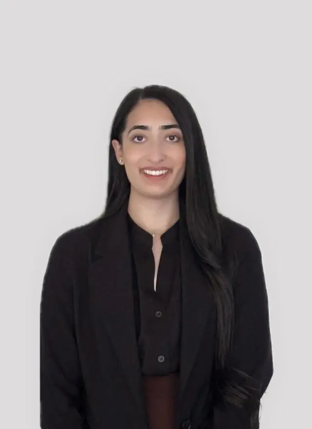A woman with long dark hair, wearing a black blazer and dark shirt, stands in front of a plain light gray background, facing the camera and smiling.