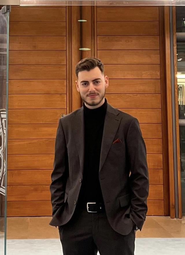 A man in a dark suit and black turtleneck stands indoors in front of wooden panel doors, with his hands in his pockets, facing the camera.
