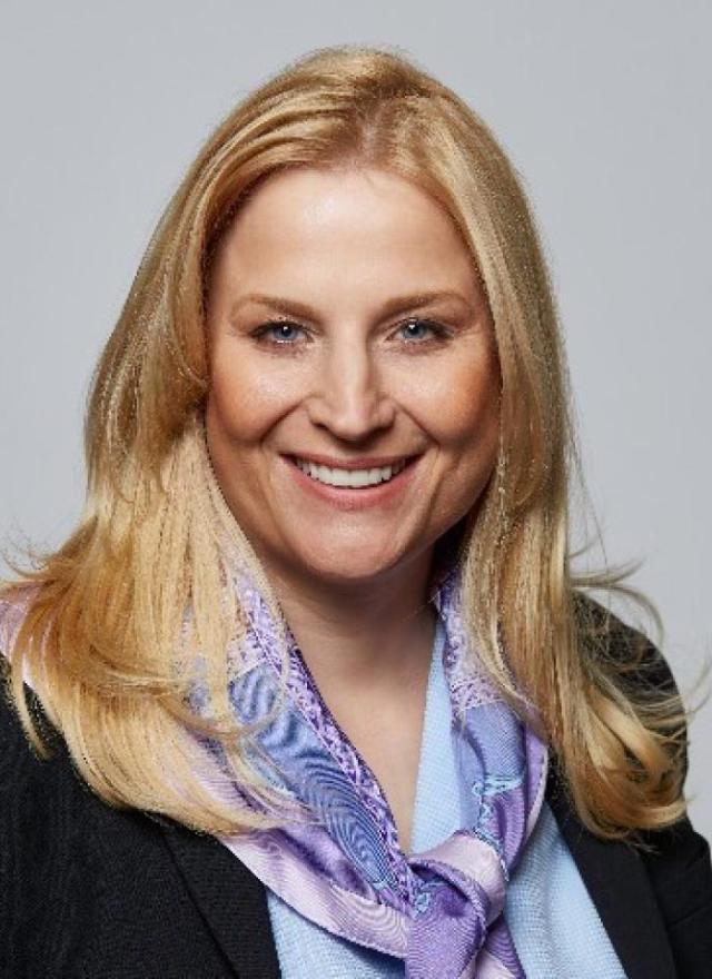 A woman with long blonde hair smiles, wearing a black blazer and a light purple patterned scarf, against a plain gray background.