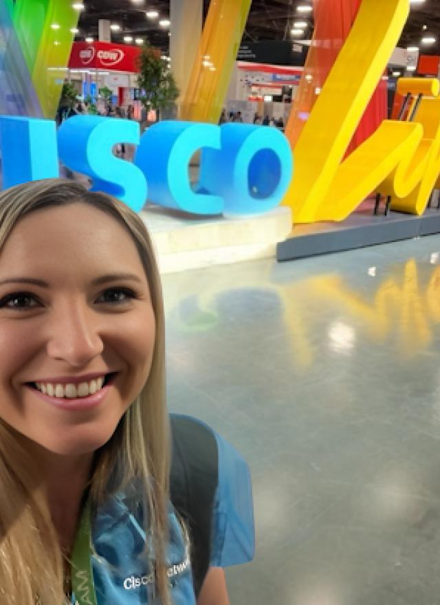 A woman takes a selfie in front of a large "CISCO Live" sign at a convention or expo, with colorful decorations and other attendees in the background.