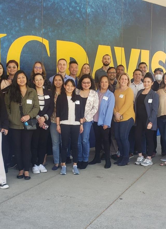 A group of people pose for a photo in front of a blue and yellow UC Davis wall on a sidewalk.