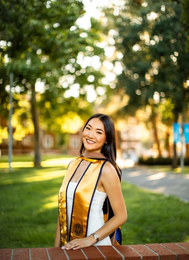 A young woman in a graduation stole and white dress stands outdoors by a brick wall, smiling, with trees and sunlight in the background.