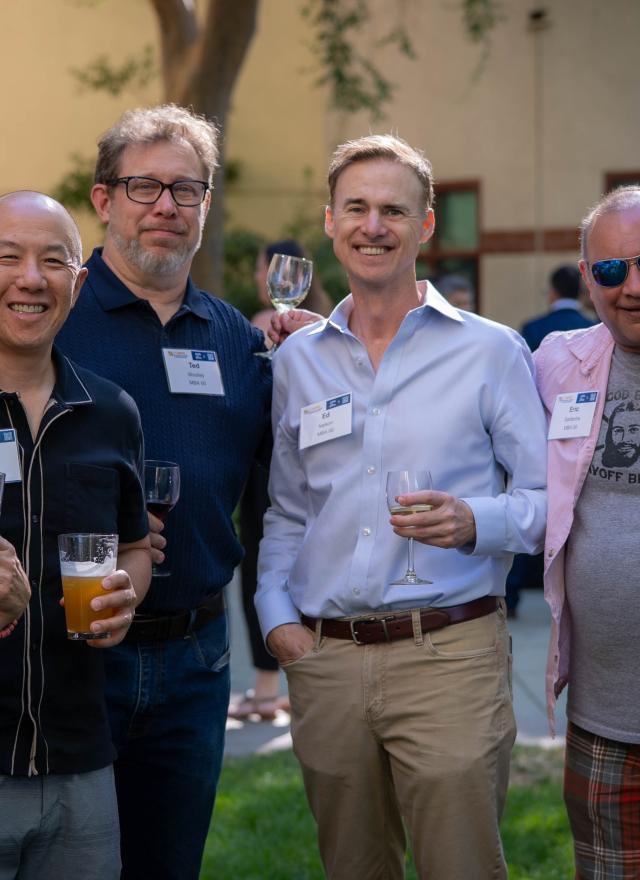 A group of six adults, wearing name tags and holding drinks, pose together and smile at an outdoor social gathering.