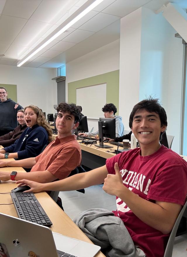 A group of students sit at desks with computers in a classroom, smiling at the camera; an instructor stands at the back of the room.
