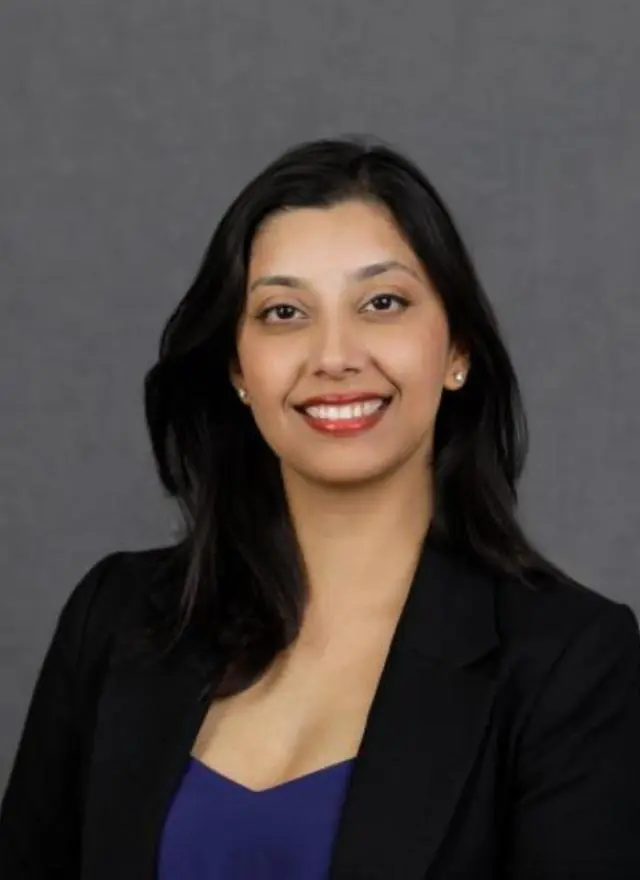 A woman with long dark hair wearing a black blazer and blue top smiles in front of a plain gray background.