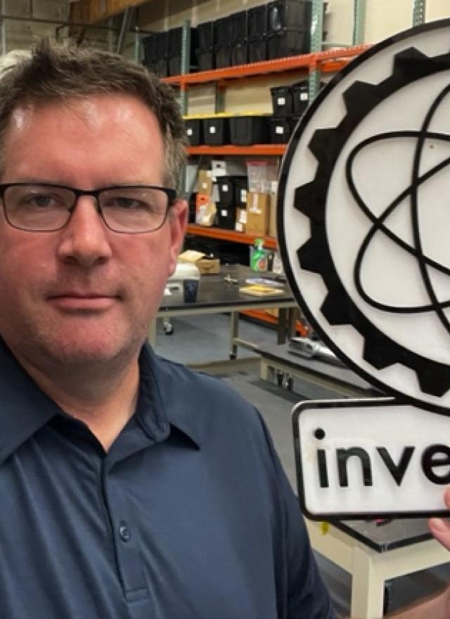 A man in a workshop holds a large sign with an atomic symbol and the word “inventopia.” Shelves and tools are visible in the background.
