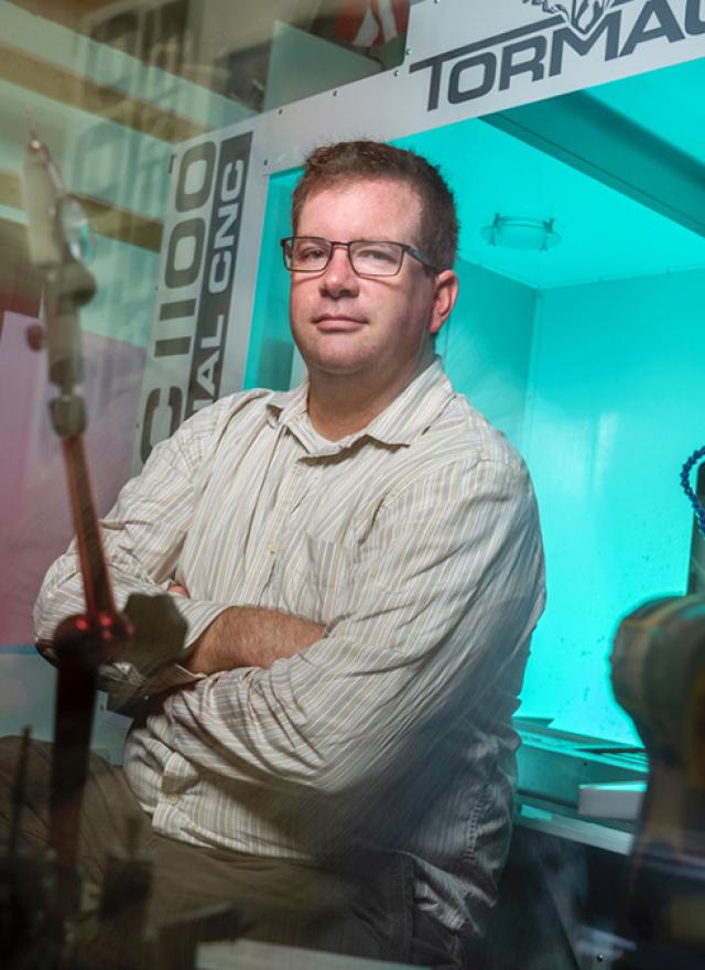 A man wearing glasses and a striped shirt sits with arms crossed in front of a Tormach CNC machine in a workshop.