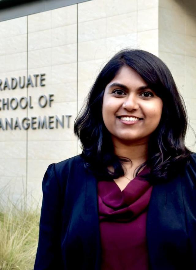 A woman in business attire stands smiling in front of a building with a sign that reads "Graduate School of Management.