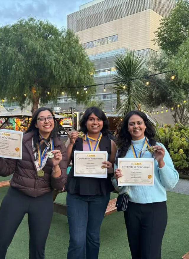 Three women stand outdoors holding certificates and wearing medals, smiling at the camera. There are picnic tables, plants, and a building in the background.