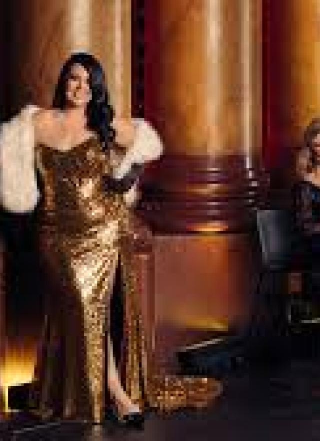A woman in a gold sequin gown stands next to a column, while another woman sits nearby playing a cello.
