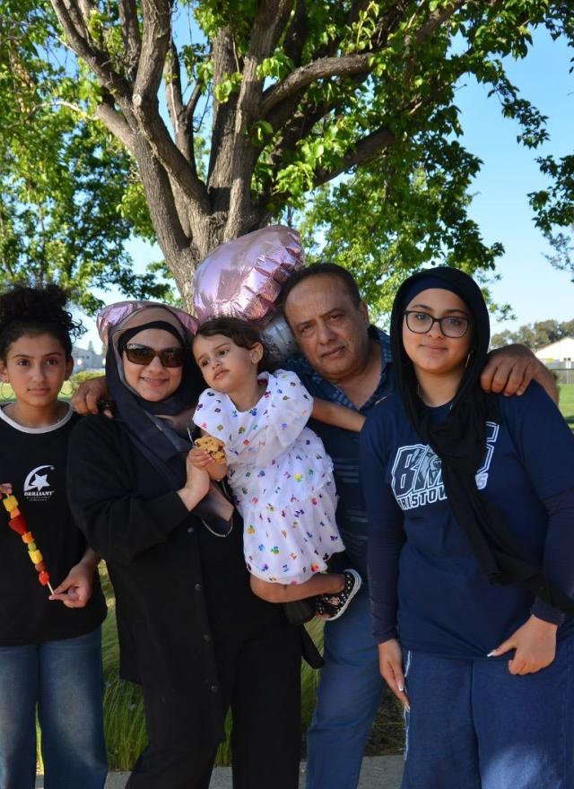 A family of five poses together outdoors in a park on a sunny day, with trees, grass, and houses in the background. One member holds heart-shaped balloons.