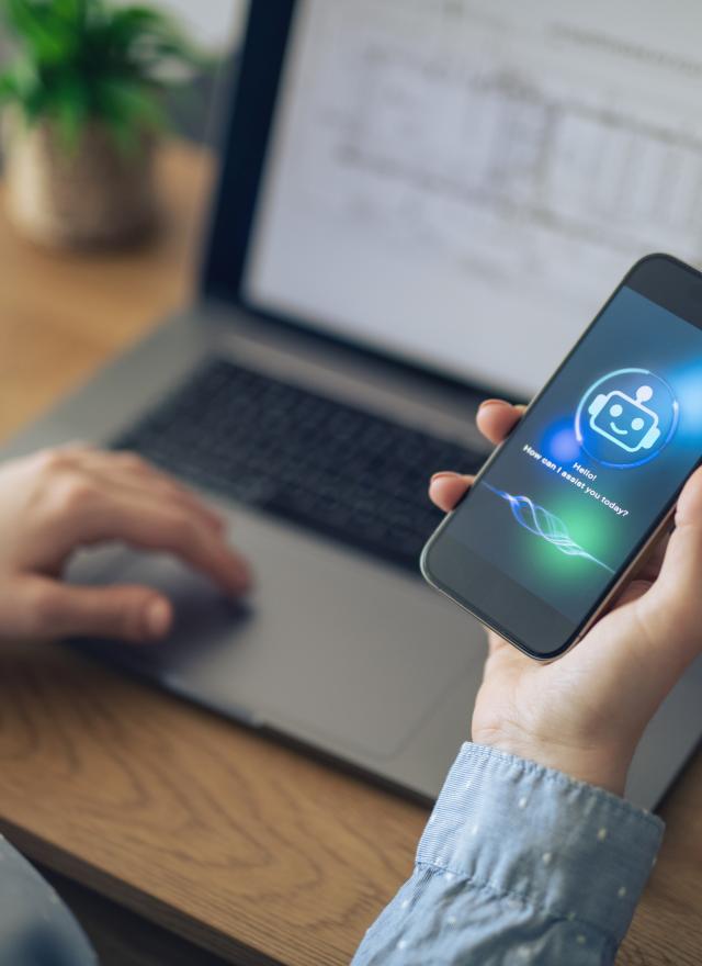 Person holding a smartphone displaying a digital assistant app icon, sitting in front of an open laptop on a desk.