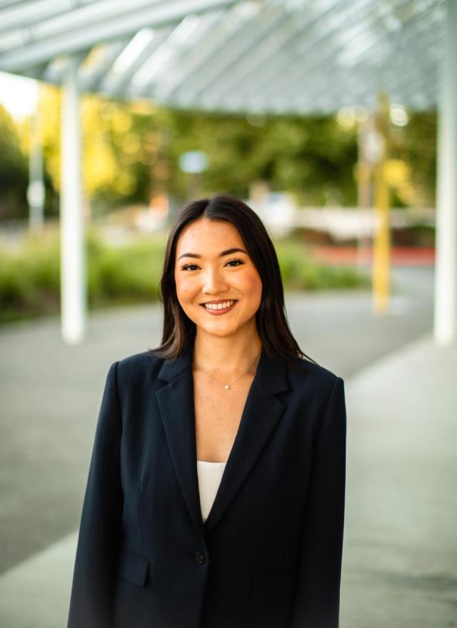 A woman in a dark blazer stands outdoors, smiling at the camera, with a blurred background of greenery and architectural structures.