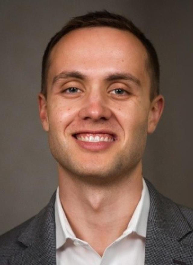 A man with short brown hair wearing a gray suit jacket and white shirt smiles at the camera against a plain background.