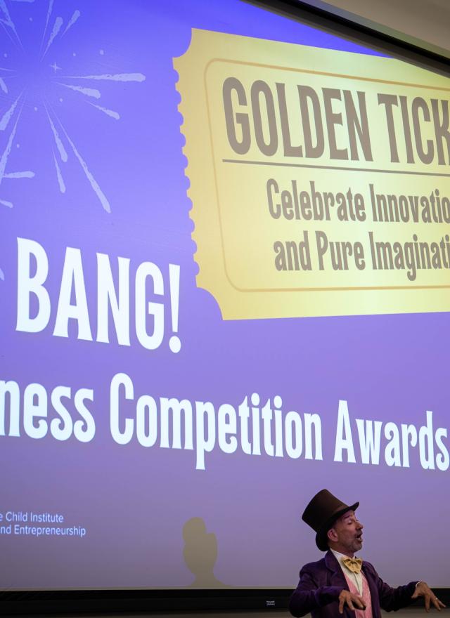 A person in a Willy Wonka costume stands in front of a projection reading "BIG BANG! Business Competition Awards" and "GOLDEN TICKET Celebrate Innovation and Pure Imagination.