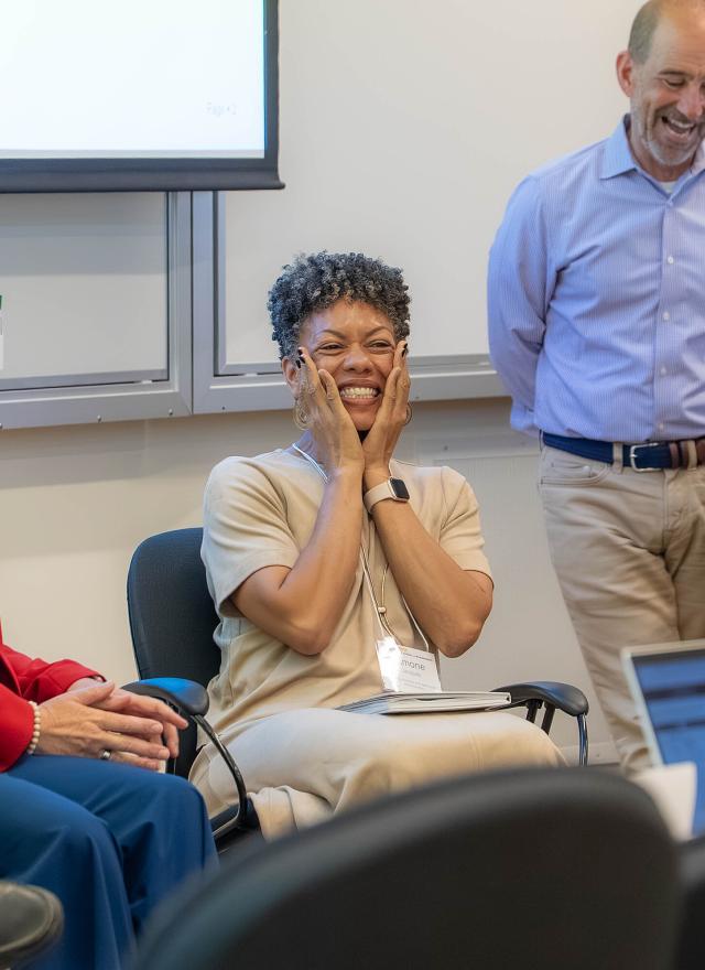 Three people sit and stand at the front of a classroom; the woman in the center smiles with hands on her cheeks, while the others look in her direction and smile.