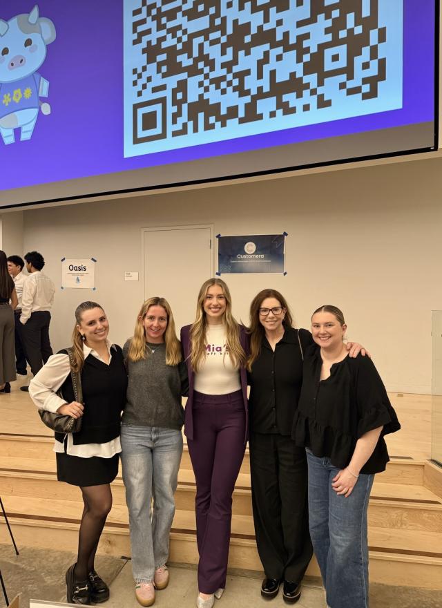 Five women stand together smiling in front of a presentation screen displaying a large QR code and a cartoon pig.