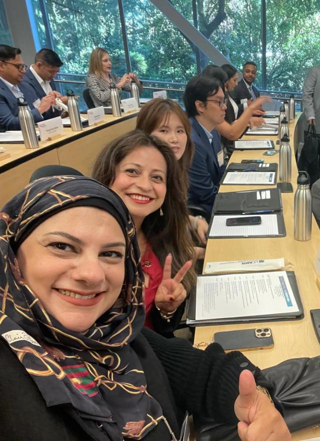 People sit in a modern classroom with large windows, facing forward. Some smile and pose for a photo, while others talk or look at materials on the desks.
