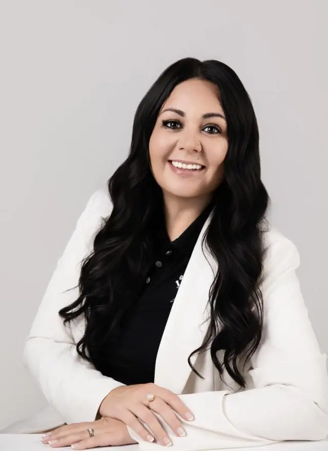 A woman with long dark hair, wearing a white blazer over a black top, is seated and smiling at the camera against a plain light background.